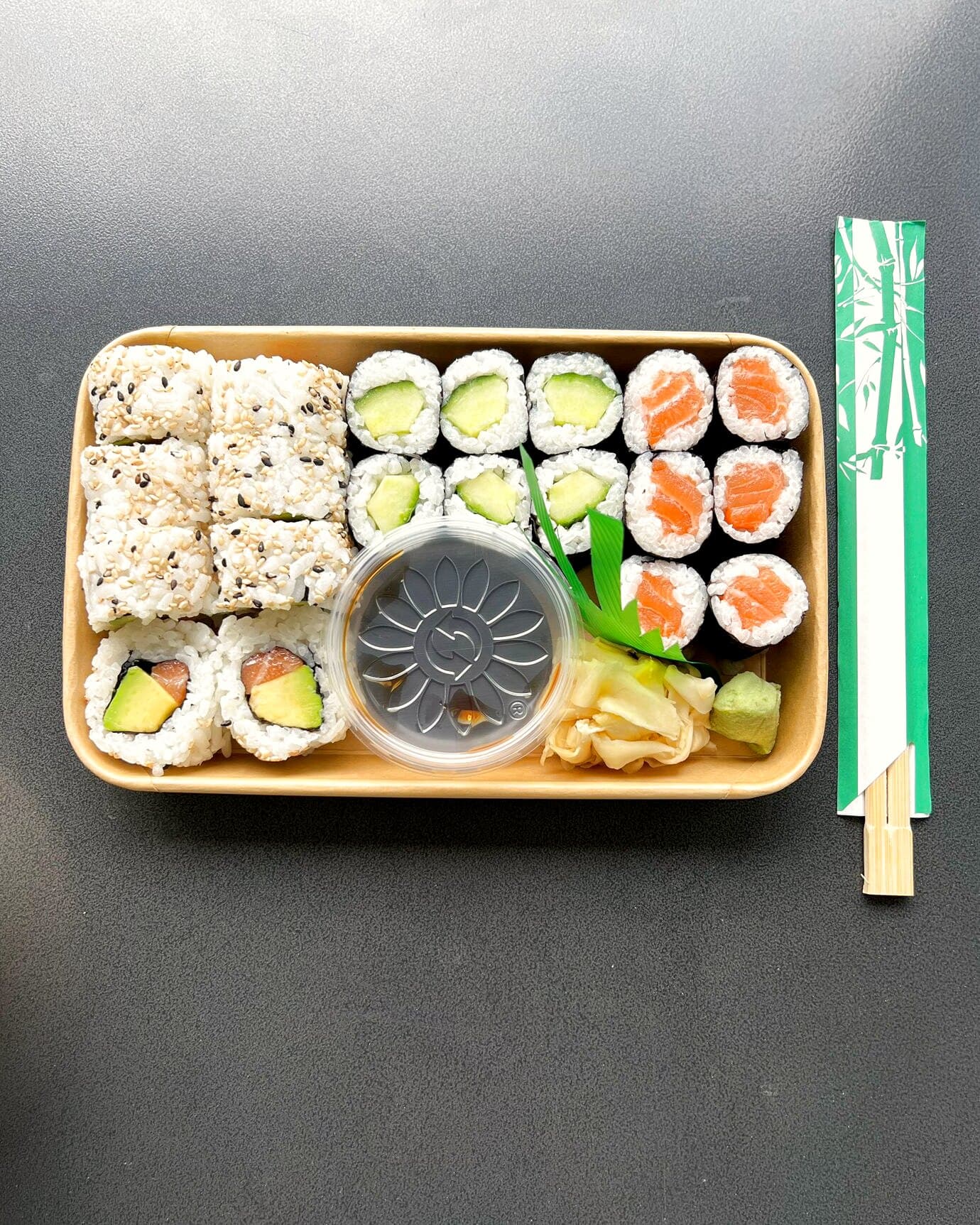 Sushi-Box von oben mit Maki und Inside-Out-Rollen, daneben Stäbchen, auf dunklem Tisch